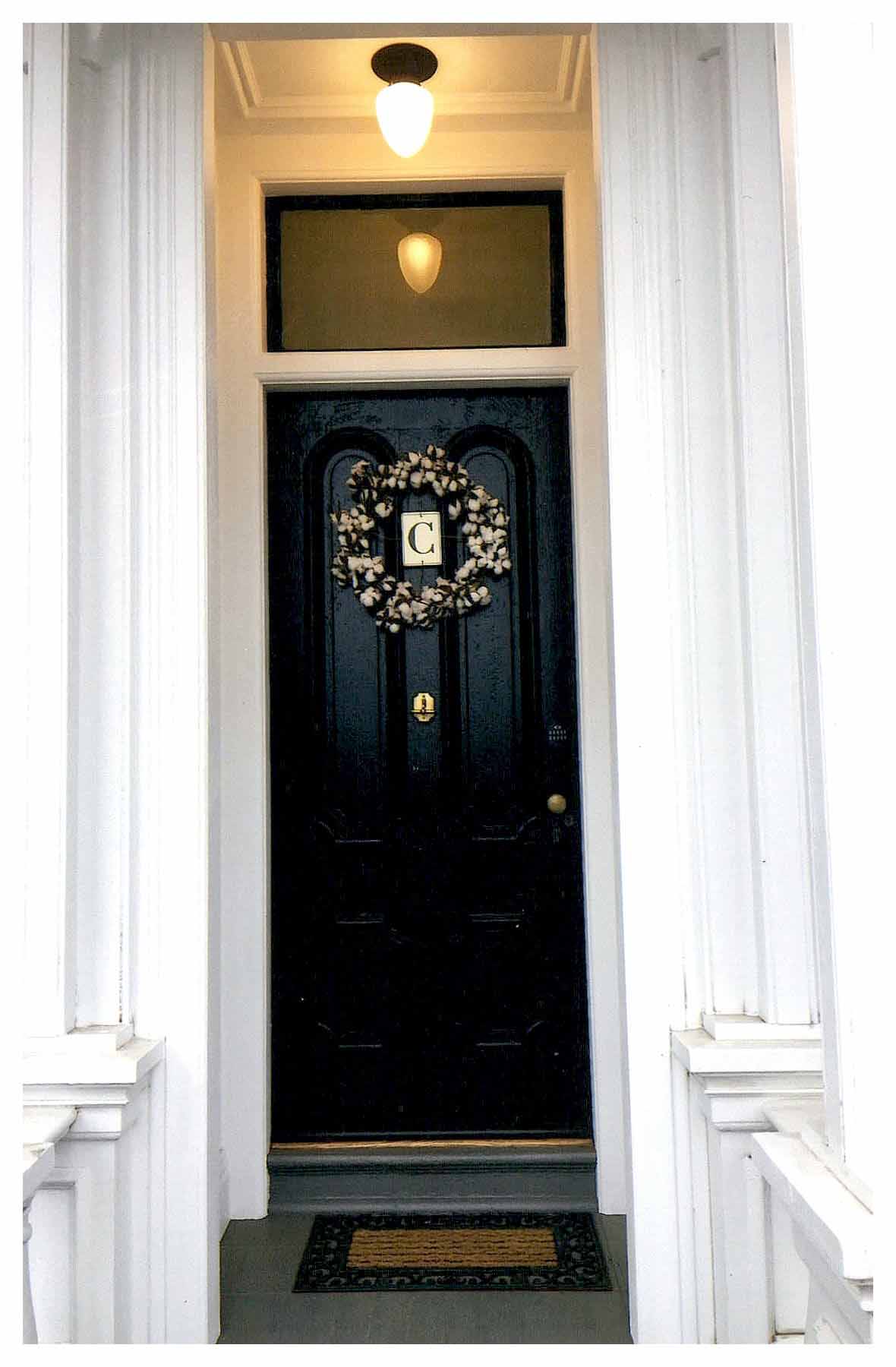 After: close-up view of restored front entrance with black painted paneled door and white flower wreath.
