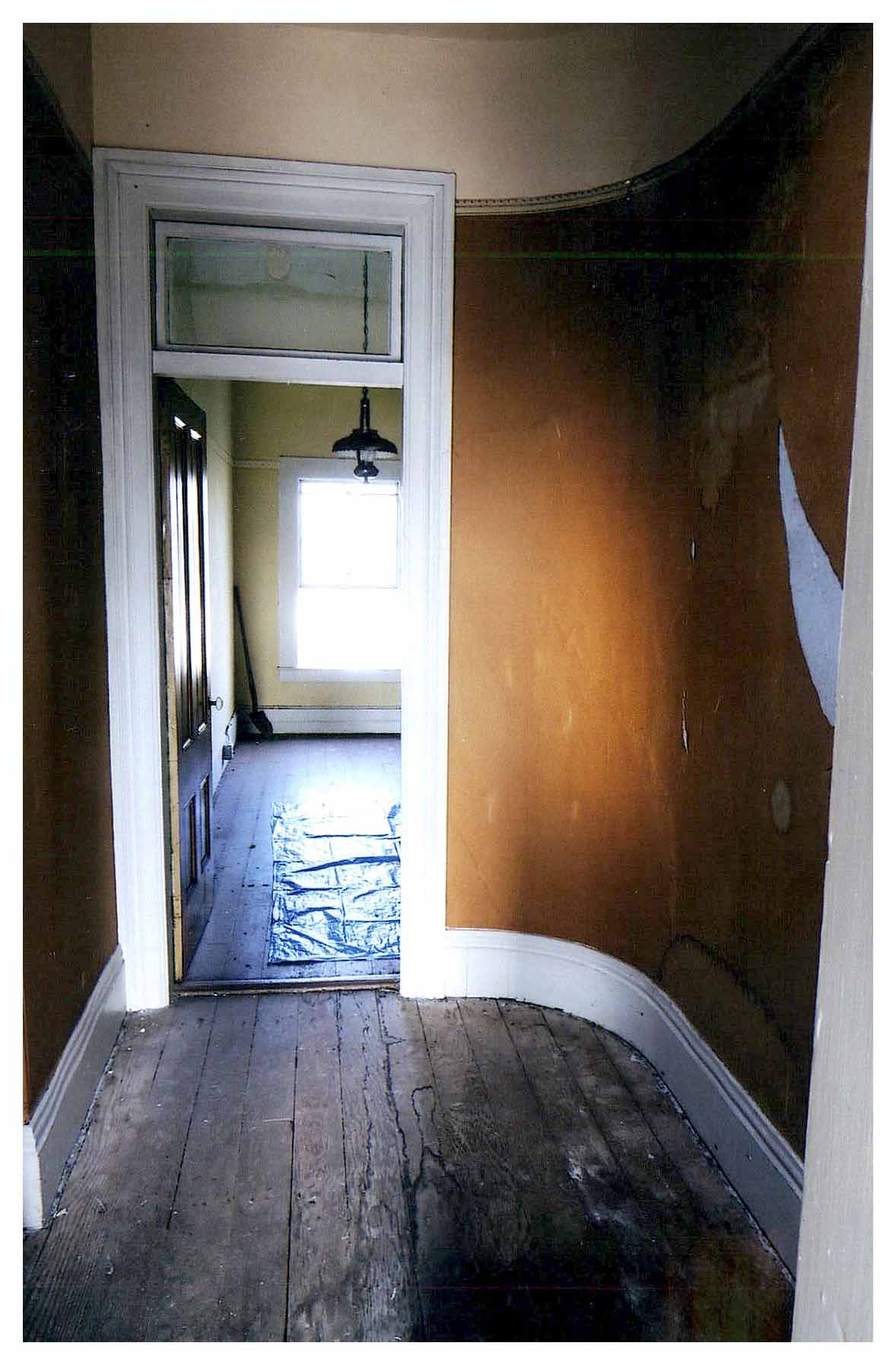 Before: same view of hallway and bedroom door with transom with unfinished wood floors and dark painted walls.