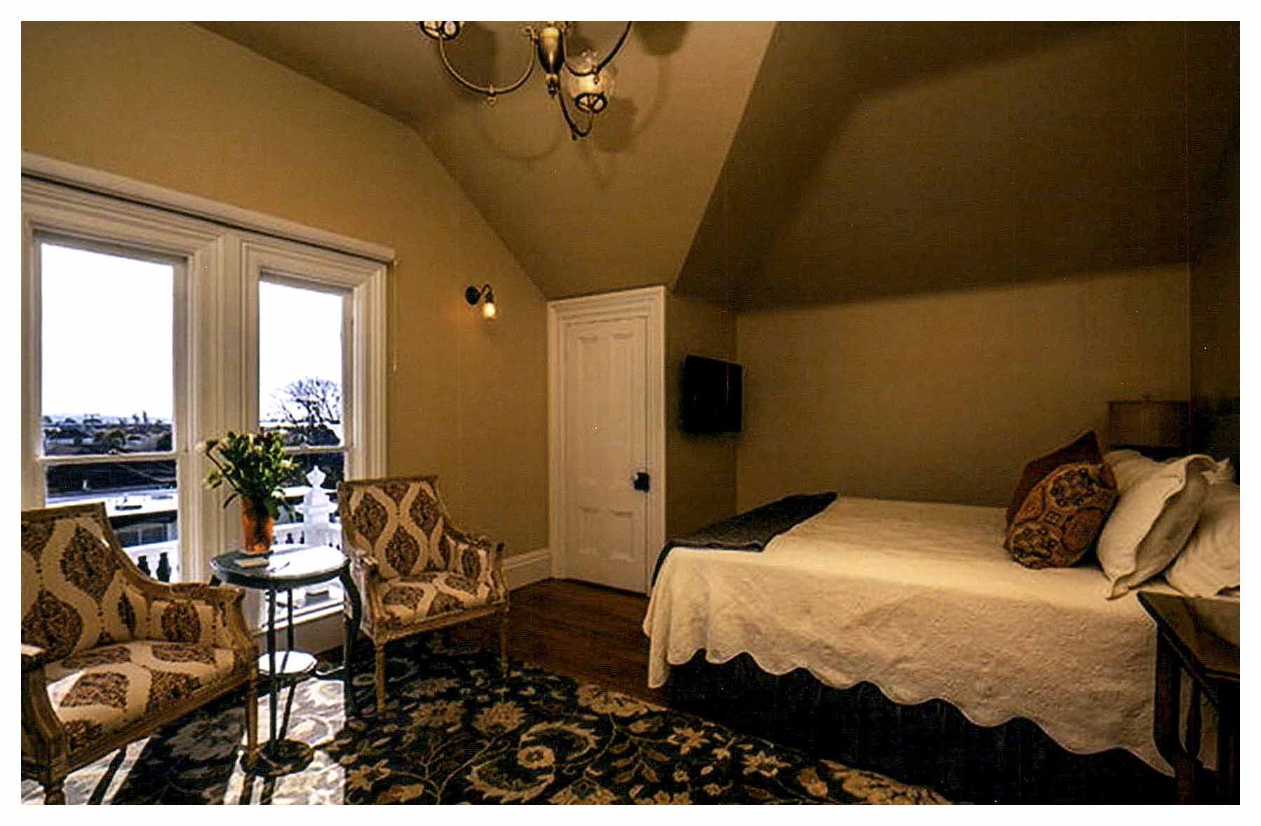 After: bedroom with mirrored plan to bedroom above including a closet under a hipped ceiling. Walls are painted beige. windows have low sills near the floor, providing much light.