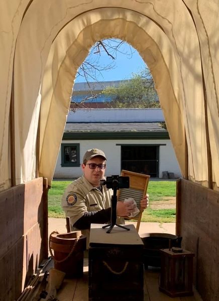 A uniformed California State Parks employee speaks into a cellphone camera set inside of a covered wagon