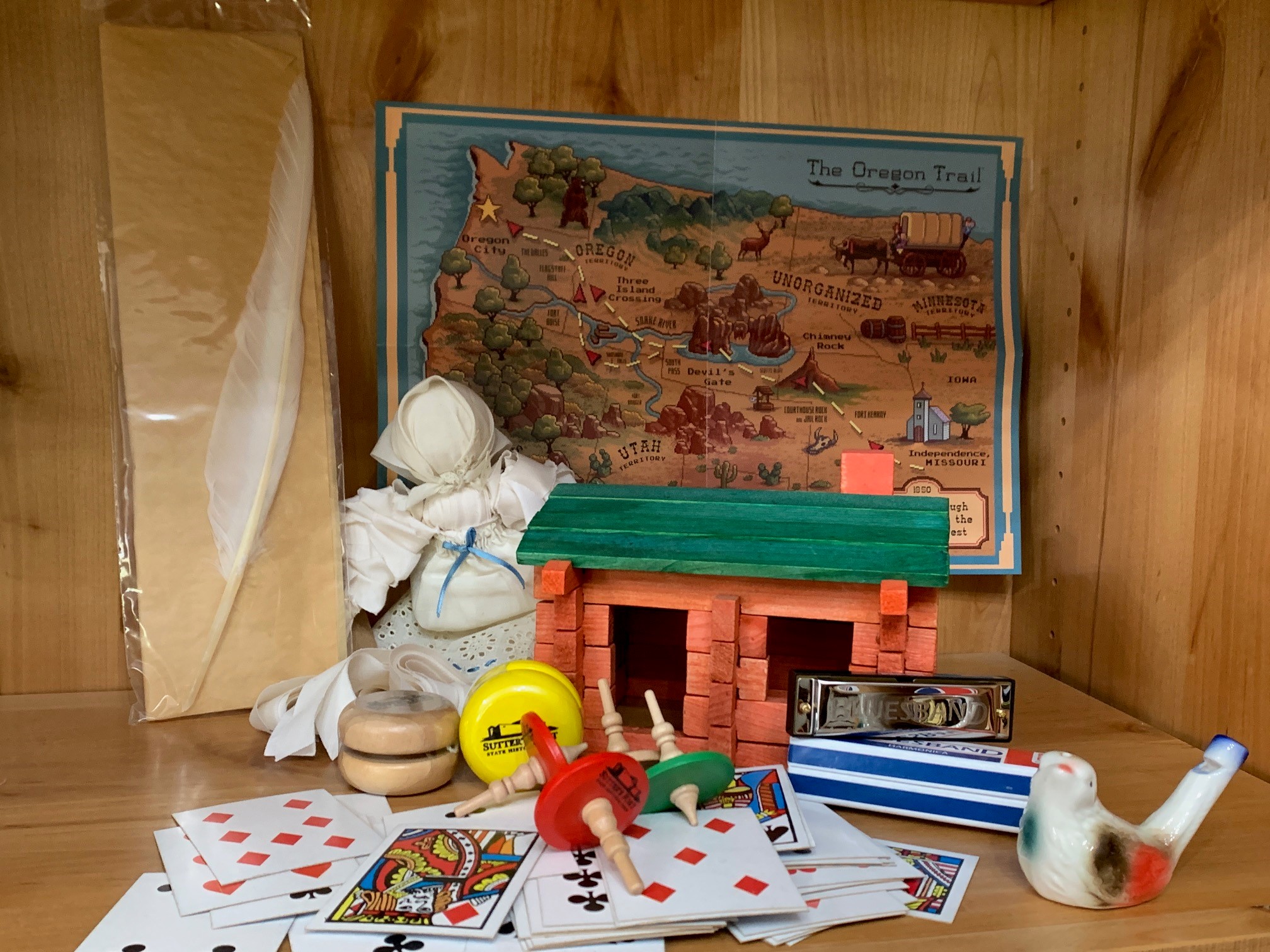retail display shelf featuring various toys: quill, rag doll, playing cards, tops, harmonica, bird whistle, yo-yos, log cabin, Oregon Trail map